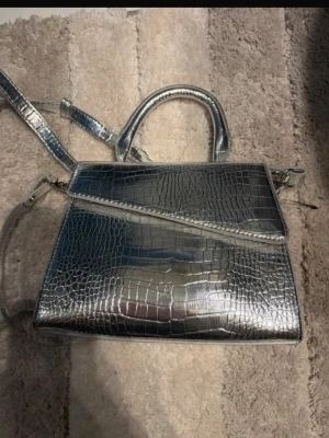 Silverfärgad handväska med krokoprägling - Snygg handväska i metallic silver med krokodilpräglat mönster. Väskan har både handtag och justerbar axelrem, samt ett snedställt lock för en trendig look. Perfekt accessoar för att lyfta vilken outfit som helst. Aldrig använd 