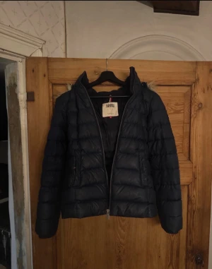 Mörkblå pufferjacka Tommy Hilfiger jeans - Snygg mörkblå pufferjacka från Tommy Hilfiger Jeans med dragkedja framtill och huva. Jackan har quiltad design och är perfekt för kalla dagar. Ett liten hål i armhålan men ingenting som märks av eller syns