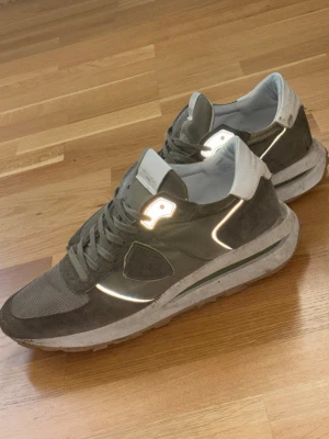 Philippe Model trpx gröna - Snygga olivgröna sneakers från Philippe Model med detaljer i mocka och mesh. Skorna har vita reflexdetaljer, vit hälkappa och en robust sula. Klassisk låg modell med snörning och logotyp på sidan. Perfekta för dig som vill ha ett par fetta men också bekväma vardagsskor och stilren design.