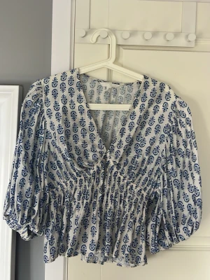 Blåvit mönstrad blus med puffärmar - Blus med blå blommönster på vit bakgrund, djup v-ringning och smockad midja. Puffiga långärmar och luftig passform. Perfekt för dig som gillar boho-stil och vill ha något unikt i garderoben.