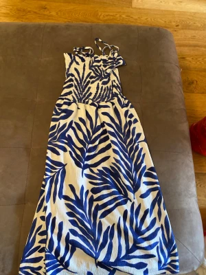 Blåvit sommarklänning med bladmönster - Säljer en snygg blåvit klänning med breda axelband och smock upptill. Klänningen har ett stort blått bladmönster på vit botten och är gjord i ett lätt, luftigt material – perfekt för varma dagar. Passformen är normal och modellen är ärmlös.