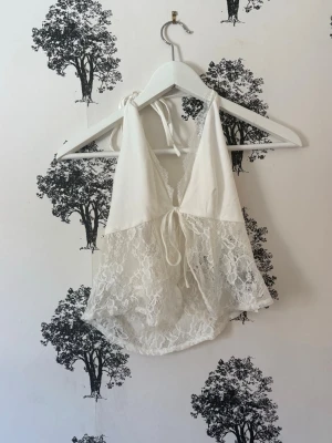 Vit spetsig linnetopp med knyt - Snygg vit linnetopp med spetsdetaljer och knyt i både nacke och rygg. Toppen är ärmlös och har en v-ringad front samt öppen rygg. Perfekt för sommarens varma dagar och ger en romantisk vibe med sitt ljusa spetsmaterial.