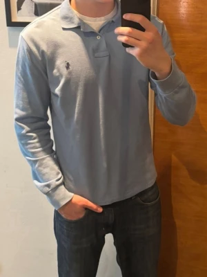 Ljusblå långärmad piké från Polo Ralph Lauren - Snygg ljusblå långärmad pikétröja från Polo Ralph Lauren med klassisk krage och två knappar framtill. Tröjan har den ikoniska loggan broderad på bröstet och är tillverkad i mjukt bomullsmaterial. Perfekt till jeans eller chinos för en clean look.