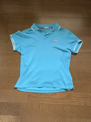Blå Moncler piké t-shirt - Tröjan är i använt men gott skick. Snygg blå piké t-shirt från Moncler med vitrandiga detaljer på krage och ärmslut. Klassisk passform med korta ärmar och knappslå framtill. Moncler-logga broderad på bröstet. Perfekt till en avslappnad men stilren look. Ha i åtanke att Moncler är slimfit.