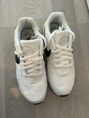 Nike vita sneakers med svart swoosh - Snygga vita Nike sneakers med svart swoosh på sidan och detaljer i neongult. Ovandelen är i mesh och syntetmaterial för skön känsla och bra andningsförmåga. Klassisk låg modell med vita skosnören och vadderad plös för extra komfort.
