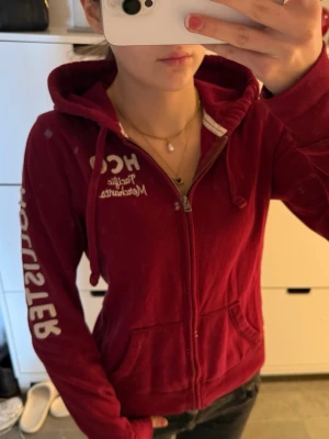 Vinröd Hollister zip up hoodie - Vintage Hollister-hoodie med dragkedja i en unik vinröd färg. Storlek L men passar bättre som en vanlig S! Mycket bra skick, förvänta dig ett litet fel på armen (sista bilden). Har den vita Hollister-loggan på bröstet och på armen. Följ för fler liknande kläder och köp bundle!