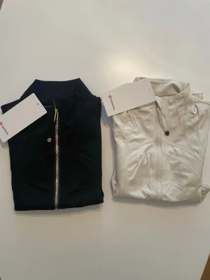 Lululemon 2 - pack - Två snygga Lululemon define jackets, en svart och en vit. Båda har hel dragkedja framtill, höga kragar och fickor med dragkedja. Västarna är ärmlösa och har en sportig, figurnära passform. Perfekta för lager på lager och stilrena detaljer.