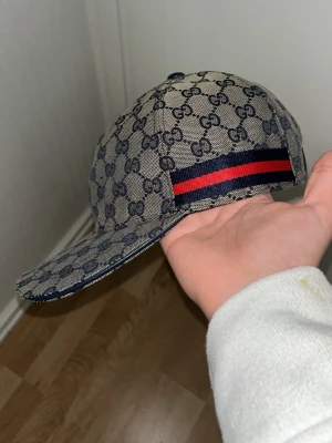 Gucci keps - Säljer en beige keps från Gucci med klassiskt blått GG-monogram över hela kepsen. På sidan finns en ikonisk röd och marinblå rand. Kepsen har böjd skärm och justerbar baksida. Materialet är canvas med exklusiv känsla, perfekt för dig som vill sticka ut.