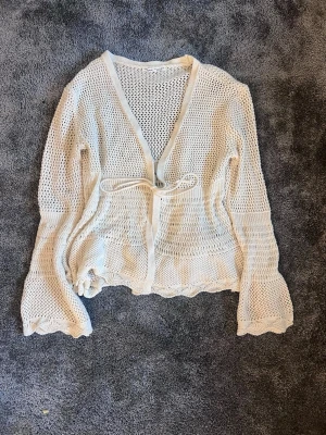 Vit virkad kofta med knytband - Snygg vit virkad kofta med v-ringning och knytband i midjan. Koftan har långa, lätt utsvängda ärmar och vågig kant nedtill. Perfekt att slänga över ett linne eller klänning för en chill look.