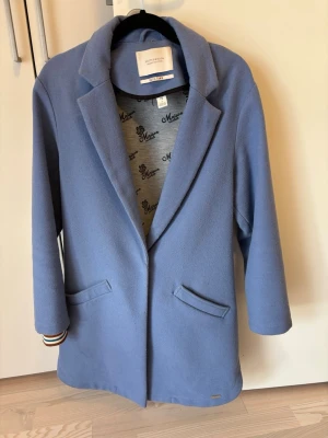 Blå kappa från Maison Scotch - Stilren blå kappa från Maison Scotch med klassisk krage och två fickor framtill. Kappan har en rak passform och är fodrad med ett mönstrat innerfoder. Perfekt för dig som vill ha en enkel men snygg look.