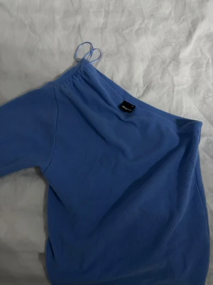 Blå oneshoulder topp från gina tricot - Säljer en blå oneshoulder topp från gina tricot med lång ärm och ribbad struktur. Toppen har en smal axelrem på ena sidan och är perfekt för dig som vill ha en trendig och unik look. Materialet känns mjukt och stretchigt.