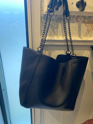 Svart shoppingväska med kedjehandtag - Stilren svart shoppingväska i konstläder med dubbla kedjehandtag i metall och rymligt innerfack. Väskan har en mjuk struktur och minimalistisk design, perfekt för dig som gillar en clean look. Invändigt finns ett mindre fack med dragkedja för småsaker.