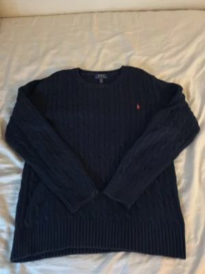 Mörkblå kabelstickad tröja Polo Ralph Lauren - Klassisk mörkblå kabelstickad tröja från Polo Ralph Lauren med rund halsringning och den ikoniska röda logotypen broderad på bröstet. Tröjan har långa ärmar och ribbade muddar vid ärmslut och nederkant. Perfekt för lager på lager under kyliga dagar.
