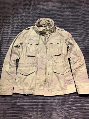 Beige camojacka från Superdry - Säljer en beige kamouflagemönstrad jacka från Superdry med fyra stora fickor framtill, hög krage och axelklaffar. Jackan har patch med brittisk flagga på ärmen och dragkedja med tryckknappar. Perfekt för dig som gillar streetwear och utilitystil.