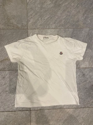 Vit t-shirt från Moncler - Klassisk vit t-shirt från Moncler med rund halsringning och liten Moncler-logga på bröstet. Tillverkad i mjuk bomull och har en enkel, stilren design med korta ärmar. Perfekt basplagg för en clean och avslappnad look.