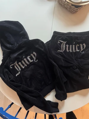 Juicy Couture svart velourset - Säljer ett ikoniskt svart velourset från Juicy Couture med strassdetaljer och texten 'Juicy' på både hoodie och byxor. Setet har en mjuk känsla, huva på tröjan och resår i midjan på byxorna. Perfekt för chill dagar hemma eller när du vill vara extra comfy.