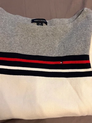 Stickad tröja Tommy Hilfiger grå/vit - Snygg stickad tröja från Tommy Hilfiger i grått och vitt med marinblå, röda och vita ränder över bröstet och ärmarna. Liten logga på bröstet. Långärmad och i mjukt material, perfekt till vardagslooken.