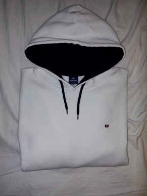 Vit hoodie från Redway - Snygg vit hoodie från Redway med svart innerfoder i huvan och svarta dragsnören. Klassisk liten Hilfiger likande logga på bröstet och känguruficka framtill. Tillverkad i mjukt bomullsmaterial, perfekt för en avslappnad stil.