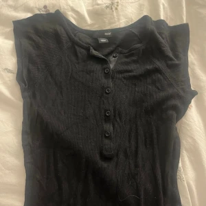 Svart ribbad topp från H&M - Svart ribbad topp från H&M med korta ärmar och knappar framtill. Toppen har en rund halsringning och är tillverkad i mjukt material som sitter snyggt på kroppen. Perfekt att styla med jeans eller kjol för en enkel look.