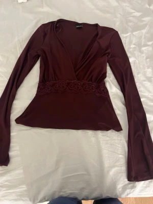 Vinröd omlottopp med spetsdetalj - Snygg vinröd omlottopp med långa ärmar och v-ringning. Toppen har en bred spetsdetalj i midjan som ger en extra touch. Tillverkad i ett mjukt, stretchigt material som sitter bekvämt på kroppen. Perfekt för dig som vill ha en stilren men ändå unik topp.