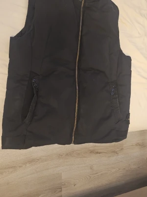 Svart dunväst med dragkedja - Snygg svart dunväst med dragkedja framtill och två fickor med knappdetaljer. Västen har en enkel och clean design, perfekt att slänga över en hoodie eller tröja. Materialet är vadderat för extra värme och komfort.