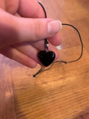 Svart hjärthalsband med snöre - Snyggt halsband med ett svart hjärta som hänge. Hjärtat är blankt och sitter på ett enkelt svart snöre. Perfekt accessoar för att ge din outfit en cool touch. Passar dig som gillar minimalistisk stil och vill ha något som sticker ut lite extra.