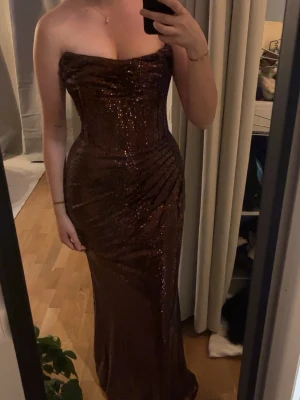 Brun glittrig axelbandslös långklänning - Säljer en brun, glittrig långklänning med axelbandslös design och figurnära passform. Klänningen har en snygg drapering över bysten och sidan, vilket ger en elegant siluett. Perfekt för dig som vill sticka ut på bal eller fest.