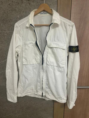 Vit overshirt från Stone Island - Vit overshirt från Stone Island med klassisk krage, två stora bröstfickor och dragkedja framtill. Ikonisk svart Stone Island-patch på vänster ärm. Tillverkad i lätt bomullsmaterial, perfekt för lager-på-lager och stilrena outfits. Jackan är i väldigt bra skick inga defekter endast lite smått smuts som ej är märkbart och säkert går att tvätta bort 
