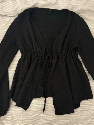 Svart omlottkofta från Gina Tricot - Svart omlottkofta från Gina Tricot med långa ärmar och knytband i midjan. Koftan har en lätt strukturerad yta och är tillverkad i ett mjukt, stretchigt material. Perfekt att slänga över en topp för en snygg lager-på-lager-look.