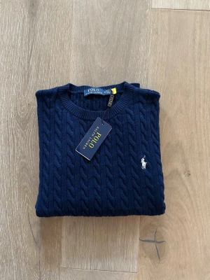 Mörkblå kabelstickad tröja Polo Ralph Lauren - Mörkblå kabelstickad tröja från Polo Ralph Lauren med rund halsringning och den klassiska vita logobroderingen på bröstet. Tröjan har långa ärmar och ribbade muddar vid ärmslut och nederkant. Tillverkad i mjuk bomull för en skön känsla. Står M men mer S!