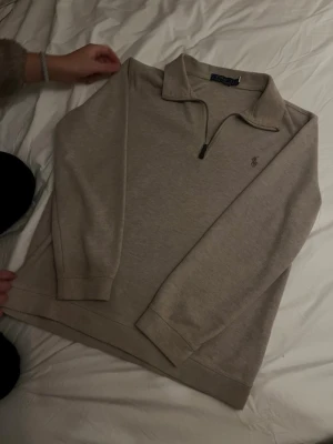 Beige stickad tröja från Ralph Lauren - Snygg beige stickad tröja från Ralph Lauren med halv dragkedja och klassisk krage. Tröjan har diskret broderad logga på bröstet och långa ärmar. Perfekt till chill vardagsstil och enkel att matcha med jeans eller kjol.