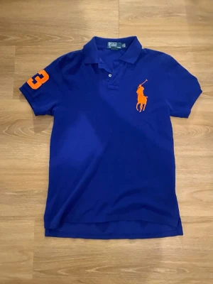 Blå pikétröja från Ralph Lauren - Klassisk blå pikétröja från Ralph Lauren med orange logga och siffra på ärmen. Slim fit-modell med krage och knappar framtill. Perfekt för en stilren och sportig look.