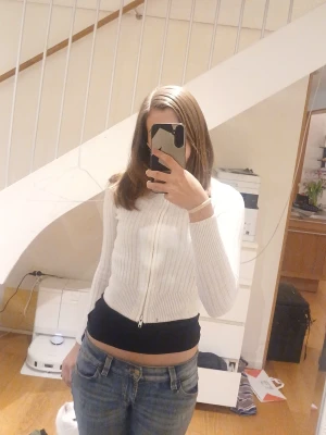 Vit ribbstickad ziptröja - Vit ribbstickad tröja med dragkedja framtill och hög krage. Modellen är croppad och har långa ärmar. Perfekt att slänga över en t-shirt eller linne för en enkel och clean look. 💕