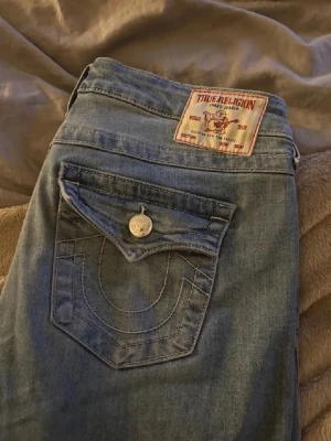Blå True Religion jeans  - Säljer ett par klassiska blå jeans från True Religion med ikonisk fickdetalj och snygga kontrastsömmar. Jeansen är utsvängda och medel höga i midjan. Säljer då dem inte används längre🤗 storlek w29 men passar M/ L. Priset går att diskutera 