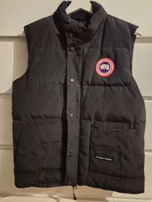 Svart dunväst från Canada Goose - Snygg svart dunväst från Canada Goose med hög krage, två stora framfickor och klassisk logotyp på bröstet. Västen har dragkedja och tryckknappar framtill samt diskret Canada Goose-märke på ena fickan. Perfekt för lager-på-lager och streetwear.