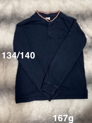 Mörkblå långärmad tröja med knappar - Mörkblå långärmad tröja med tre knappar vid halsen och en liten bröstficka. Tröjan har ribbade muddar vid ärmsluten och en kontrastfärgad kant i rött och vitt vid kragen. Perfekt för en avslappnad stil.🌸 Fint använt skick. Storlek 134/140