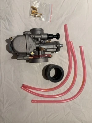 Förgasare med tillbehör till moped - Säljer en grå förgasare i metall med tillhörande rosa slangar, svart gummiadapter och ett set munstycken i plastpåse. Perfekt för dig som vill trimma eller serva din moped eller scooter. Passar flera olika modeller.