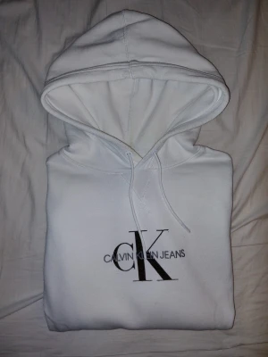 Vit hoodie från Calvin Klein Jeans - Vit hoodie med stor huva och snörning från Calvin Klein Jeans. Klassisk CK-logga i svart på bröstet. Mjuk bomullskänsla och ribbade muddar vid ärmslut och nederkant. Perfekt för en clean och stilren look.
