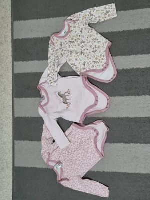 3-pack långärmade bodys i rosa toner - Säljer ett set med tre långärmade bodys i mjuk bomull. Två bodys har söta blommönster i rosa och beige, och en är ljusrosa med ett tryck av en hjort framtill. Alla har rosa kantband och tryckknappar i grenen för enkel påklädning.