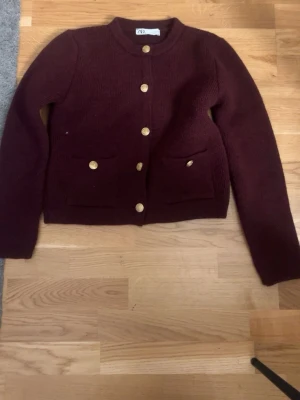Vinröd kofta från Zara med guldknappar - Mysig vinröd kofta från Zara med rund halsringning och dekorativa guldknappar framtill. Två fickor framtill ger extra stilpoäng. Perfekt till höst och vinter för en chic look.
