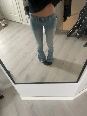 Ljusblå bootcut jeans från True Religion - Säljer ett par ljusblå bootcut jeans från True Religion med klassiska kontrastsömmar och broderade fickor bak. Jeansen har låg midja och utsvängda ben, perfekta för en avslappnad och trendig look. Ändast använda 3 gånger