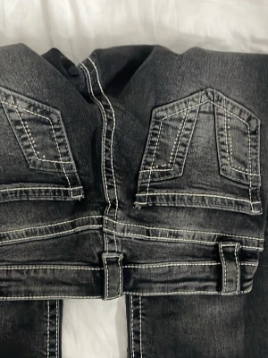 Svarta jeans med kontrastsömmar - Säljer ett par svarta jeans med tydliga vita kontrastsömmar och unika bakfickor med dubbla lock och dekorativa detaljer. Jeansen har breda hällor och en tvättad look som ger en cool och trendig känsla. Perfekta för dig som gillar streetwear och vill sticka ut.