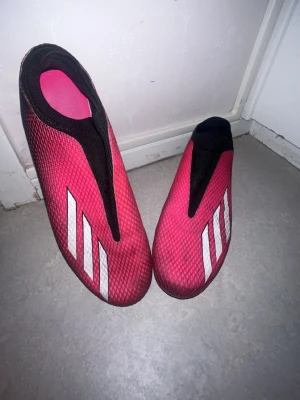 Adidas X Ghosted.3 Laceless FG - Säljer ett par rosa fotbollsskor från Adidas med klassiska vita ränder på sidan. Skorna har en strömlinjeformad design utan snörning och svart meshdetalj upptill. Perfekta för dig som vill ha stilrena och lätta skor på planen.