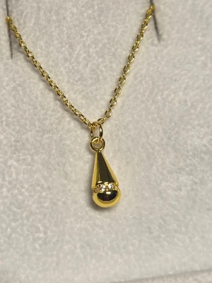 Guldfärgat halsband med konformad droppe berlock - Snyggt halsband i guldton med en konformad berlock. Berlocken har en liten rad med gnistrande stenar som detalj. Kedjan är tunn och elegant, perfekt för att ge en stilren touch till din outfit. Materialet är troligtvis metall med guldfinish.