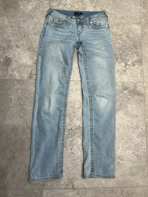 Ljusblå raka jeans från True Religion - Säljer ett par ljusblå jeans från True Religion med raka ben och klassiska fem fickor. Jeansen har snygga kontrastsömmar och ikoniska broderade bakfickor. Tillverkade i mjuk denim med en avslappnad passform. Perfekta för en casual look. De har små defekter som syns i bild och använda ett fåtal gånger! Storlek 23