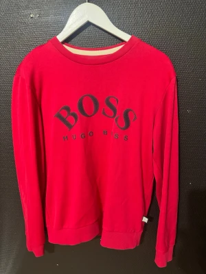 Röd sweatshirt från Hugo Boss - Riktigt snygg röd sweatshirt från Hugo Boss med stor svart logga på bröstet. Tröjan har rund halsringning, långa ärmar och ribbade muddar vid ärmslut och nederkant. Perfekt för en avslappnad och trendig look.
