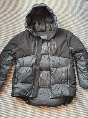 Sail Racing - Svart junior dunjacka från Sail Racing - Snygg svart junior (strl 160) dunjacka från Sail Racing med huva och dragkedja framtill. Jackan har en modern, puffig design med flera fickor och logga på bröstet. Perfekt för kalla dagar och håller dig varm med sitt dunfoder. Stilsäker och funktionell jacka för vintern. Nypris ca 1 700kr, säljes för 1 250kr. 