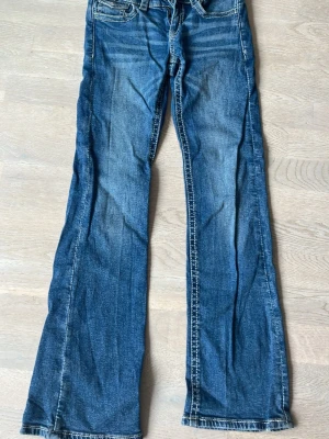 Blå bootcut jeans med detaljer - Säljer ett par blå bootcut jeans med slitningar och snygga detaljer på bakfickorna. Jeansen har klassisk femficksmodell och kontrastsömmar. Perfekta för dig som gillar en avslappnad men trendig look.