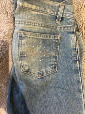 Ljusblå bootcut jeans från Dolly Jeans - Säljer ett par ljusblå bootcut jeans från Dolly Jeans med snygg slitning på benen och dekorativa detaljer på bakfickan. Klassisk femficksmodell med broderad logga och små strasstenar på fickan. Perfekta för en avslappnad och trendig look.