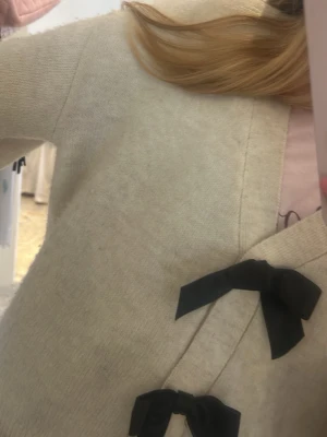Krämvit kofta med svarta rosetter - Söt krämvit kofta med två stora svarta rosetter framtill. Koftan har v-ringning och ribbade muddar vid ärmsluten. Perfekt för dig som gillar en gullig och stilren look. Materialet ser mjukt och bekvämt ut.